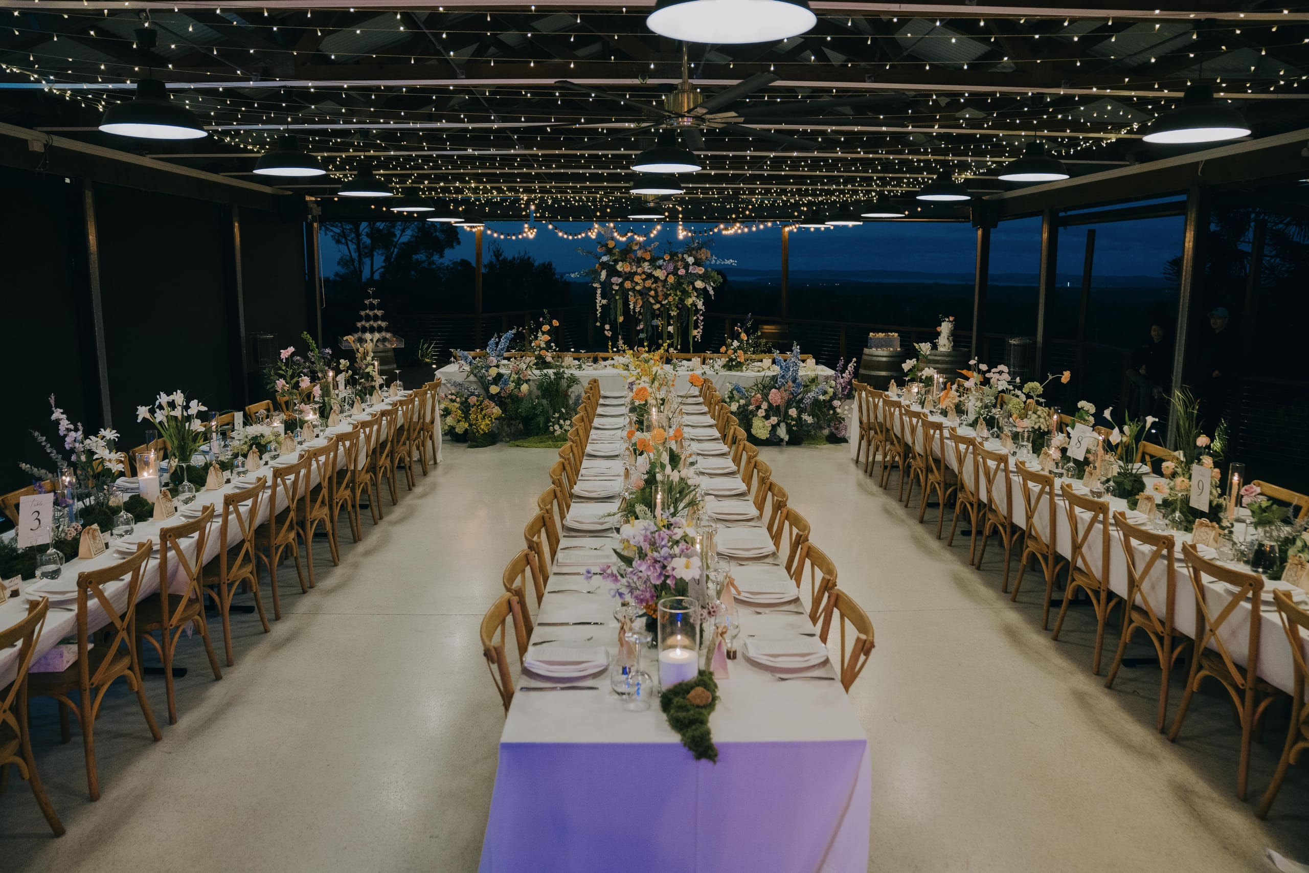 A long-table reception beneath festoon lights, with florals along every table